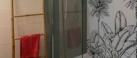 Een moderne badkamer met een glazen douchecabine, een bamboe ladder met een rode handdoek, en een wanddecoratie van tropische planten.