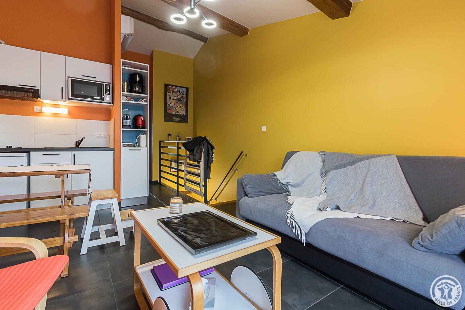 A compact living space with a kitchenette, a gray sofa, and a wooden table with a tablet.