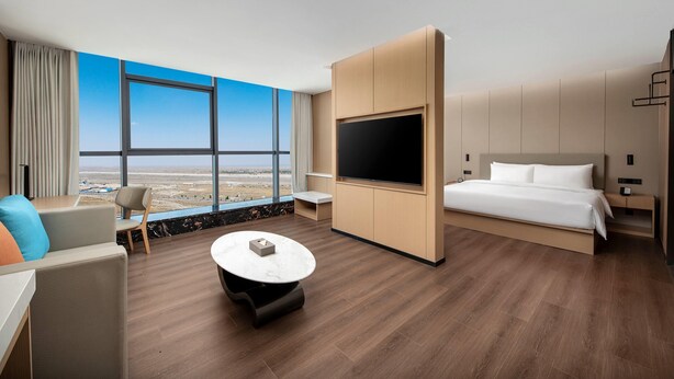 A modern hotel room with a large window, a flat-screen TV, a bed, a desk, and a round coffee table. - Ramada By Wyndham Khorgos Free Trade Zone (Huocheng County)