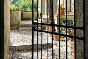 Blick durch ein vergittertes Fenster auf einen Innenhof mit Stufen, die zu einer Tür führen, umgeben von Kies und Topfpflanzen.