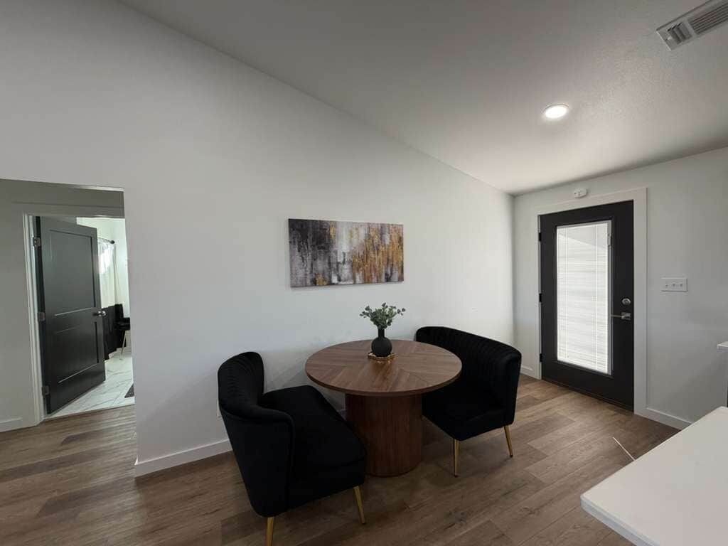 A modern room with a round wooden table, two black chairs, a painting on the wall, and a door with blinds.
