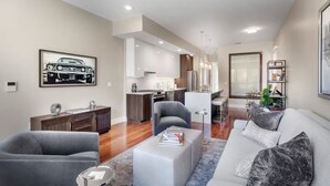 A modern living room with a grey sofa, a coffee table, and a grey armchair. There is a kitchen in the background with white cabinets and a dining area.