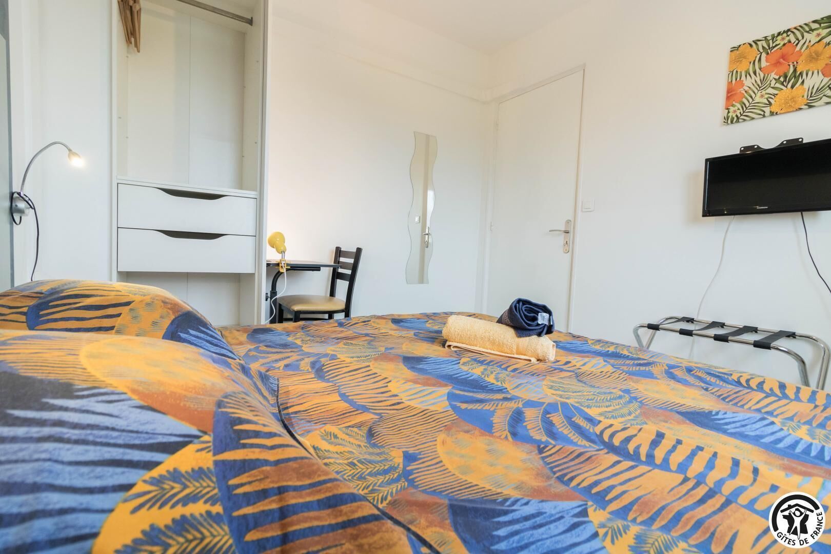 A hotel room with a bed featuring a tropical leaf pattern, a desk with a chair, a TV mounted on the wall, and a wardrobe.