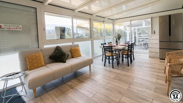 Una sala de estar moderna con un sofá, una mesa de comedor y sillas. Hay una zona de cocina con un refrigerador y una ventana con vistas a un jardín.