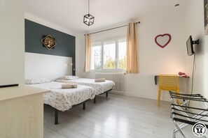 Une chambre avec deux lits, un bureau, un radiateur, une fenêtre avec des rideaux, une chaise jaune et un objet décoratif en forme de cœur accroché au mur.