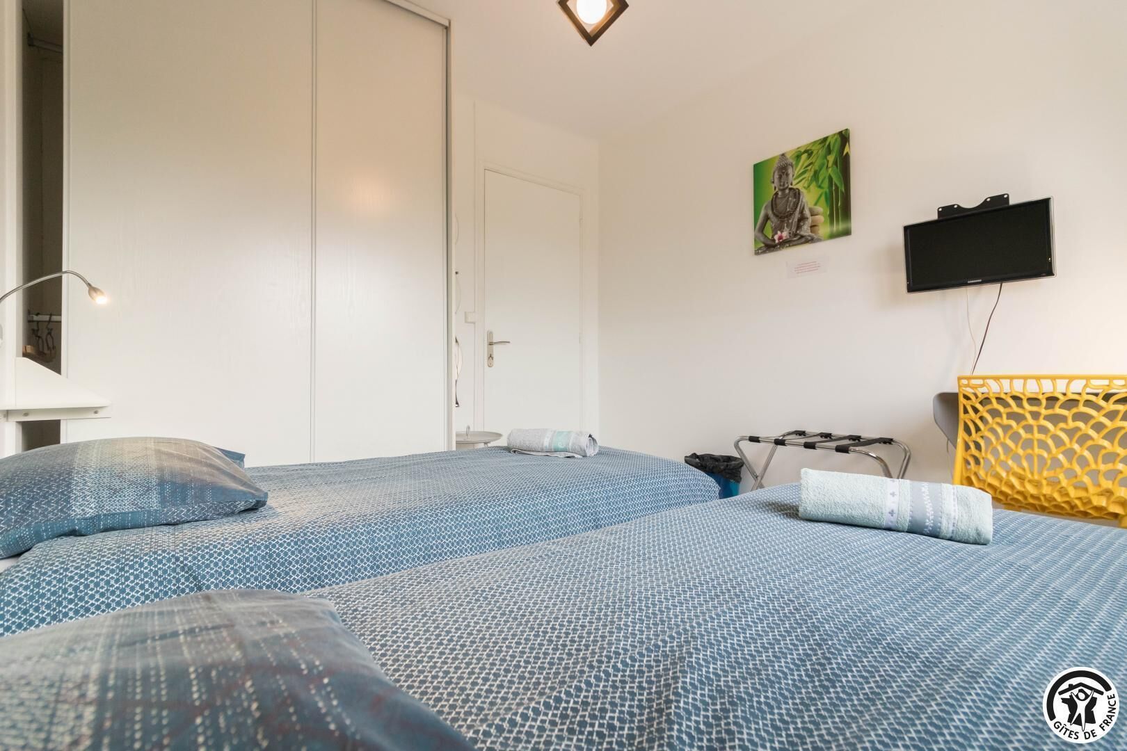 Une chambre d’hôtel avec deux lits, une télévision, un tableau et une tête de lit décorative jaune.