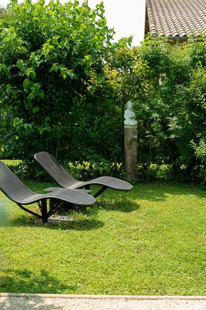Deux fauteuils de jardin sont placés dans un espace verdoyant, entouré d’une végétation luxuriante, et on aperçoit en arrière-plan une structure avec un toit en tuiles.