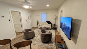 Una sala de estar con un sofá color crema, dos sillones, una mesita de centro, un televisor en la pared y una estantería con libros.