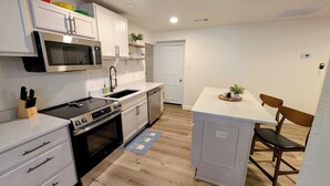 Cocina compacta con armarios blancos, un microondas, estufa y pileta.  Un pequeño comedor con una silla y una mesa.