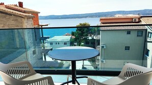 A balcony with a glass barrier, two white plastic chairs, a round table, and a view of a coastal town. - Apartment Azure Suite in Lokva Rogoznica (Lokva Rogoznica)