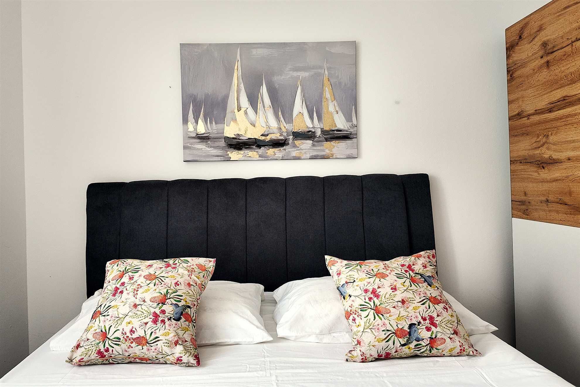 A bed with two floral pillows, a dark headboard, and a painting of sailboats on the wall.