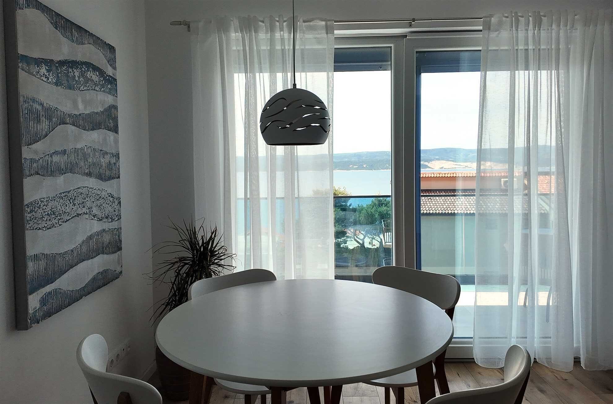 Ein Essbereich mit rundem Tisch und Stühlen, einer Hängeleuchte, einem großen Fenster mit durchsichtigen Vorhängen und Blick auf eine Landschaft.