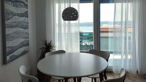 A dining area with a round table and chairs, a hanging light fixture, a large window with sheer curtains, and a view of a landscape. - Apartment Azure Suite in Lokva Rogoznica (Lokva Rogoznica)