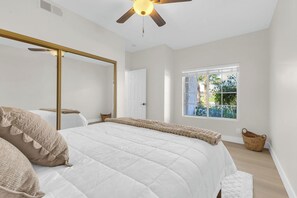 A bedroom with a large bed, a ceiling fan, a window with a view of greenery, and a white door. - Golf Course l Pools l Views l Tennis & Pickleball (Palm Desert)
