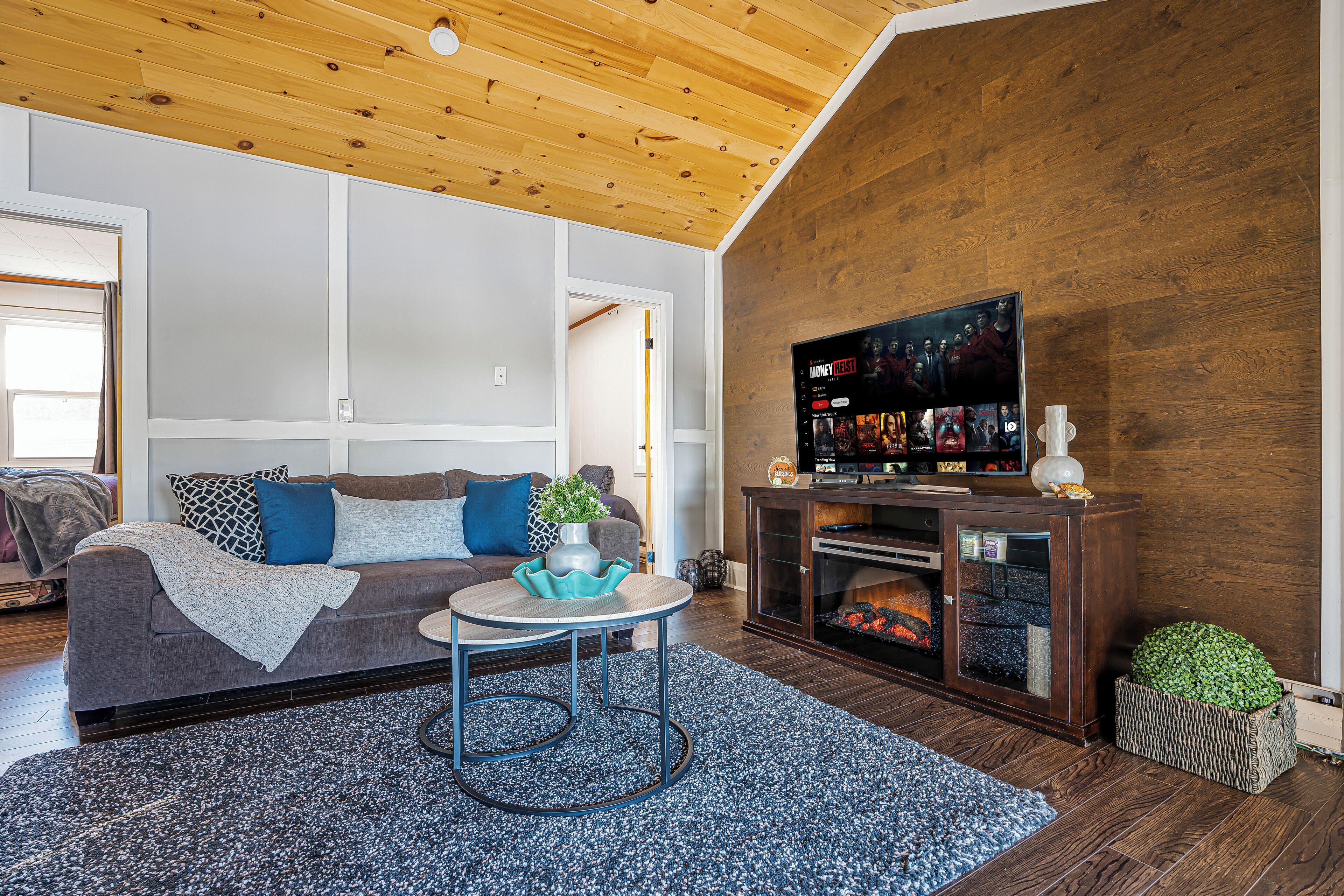 A modern living room with a wooden ceiling, a grey sofa with blue cushions, a round coffee table, and a television on a wooden stand.