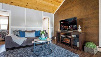 A modern living room with a wooden ceiling, a grey sofa with blue cushions, a round coffee table, and a television on a wooden stand.
