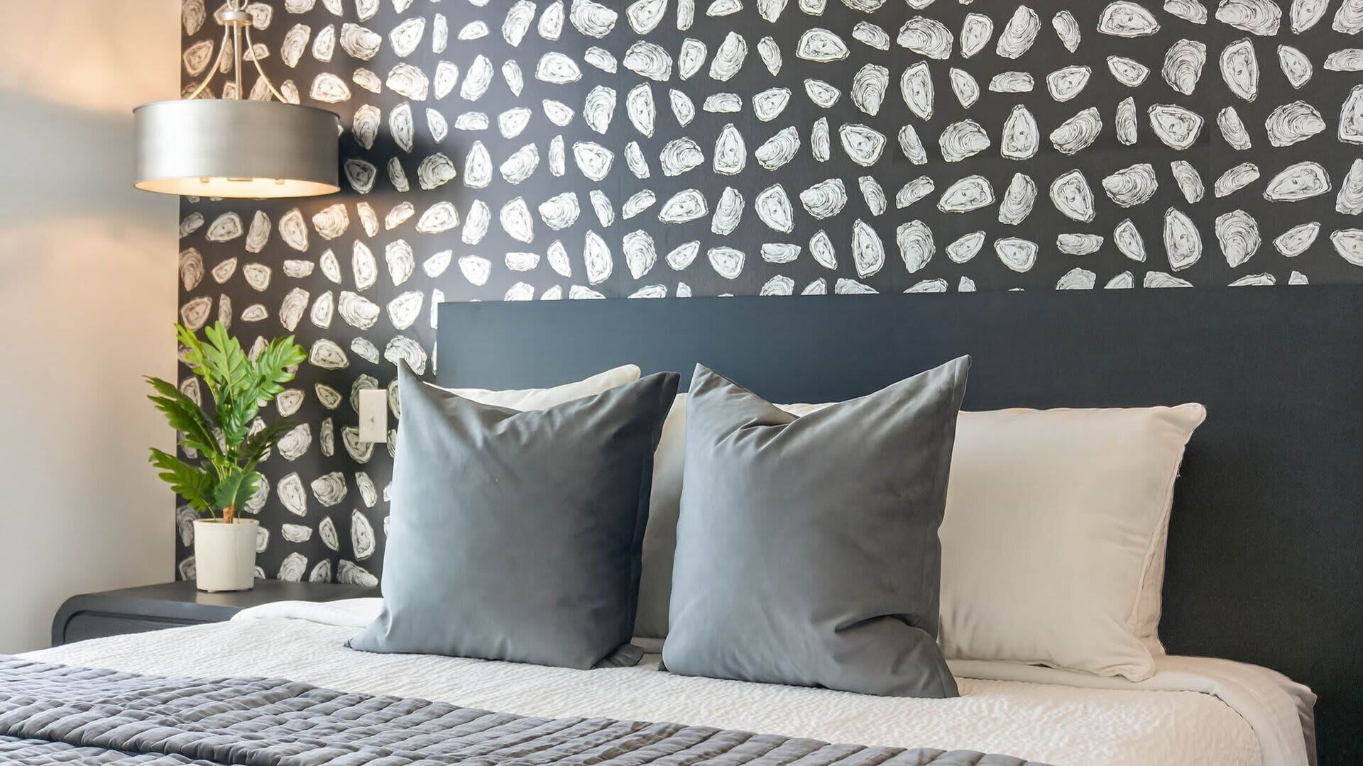 Ein Bett mit grauen und weißen Kissen, ein dunkles Kopfteil, eine Zimmerpflanze am Nachttisch und eine Wand mit Blattmuster.