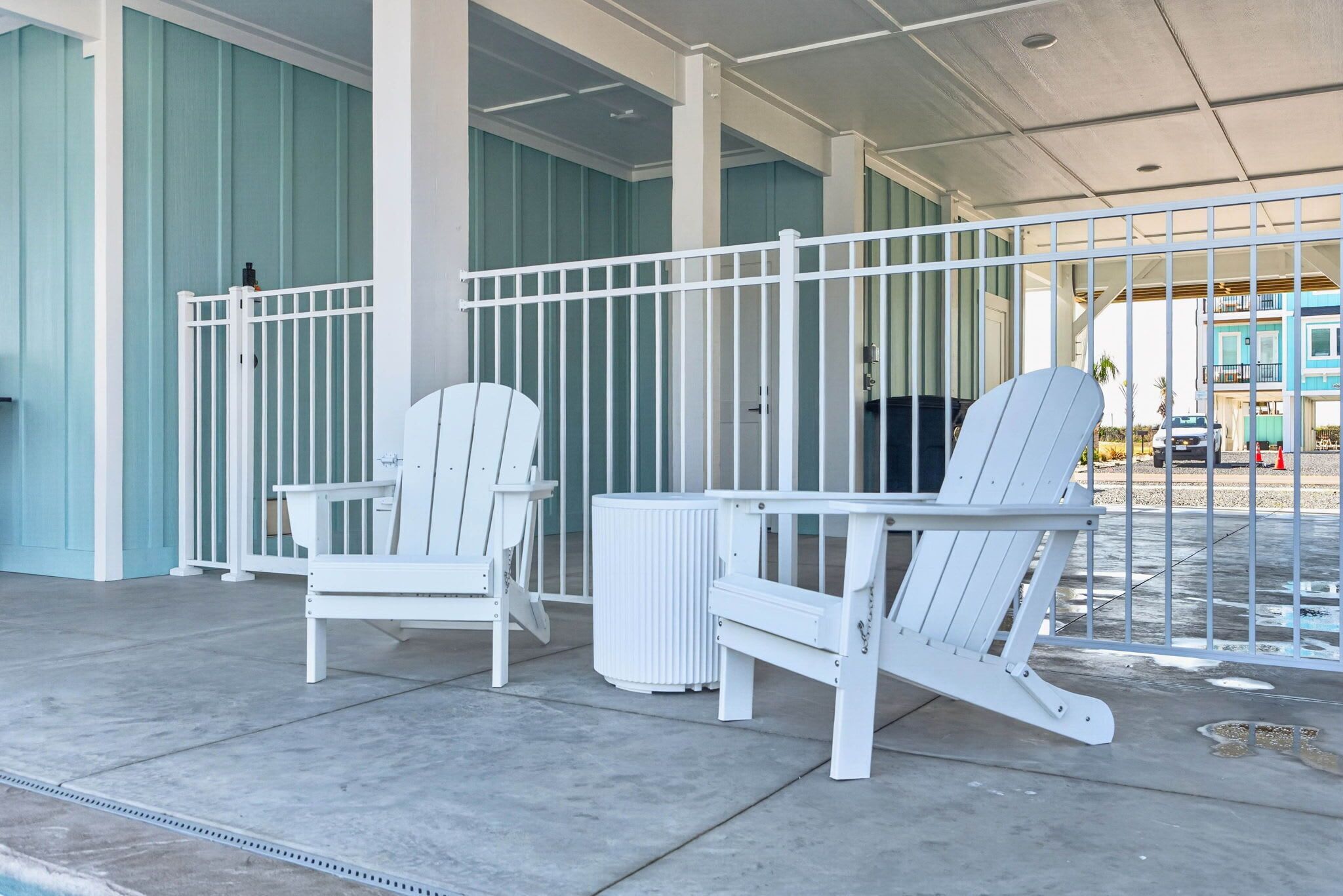 A covered outdoor seating area with white Adirondack chairs and a white table, a white railing, and a view of a building in the background.