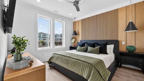 Un dormitorio moderno con cama, mesitas de noche, un escritorio y vistas a casas a través de amplias ventanas.