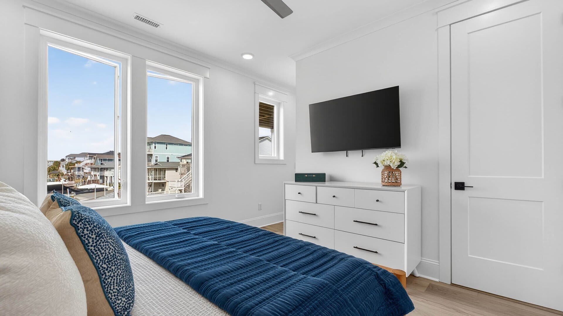 A bedroom with a bed, a dresser, a TV, and a window with a view of buildings.