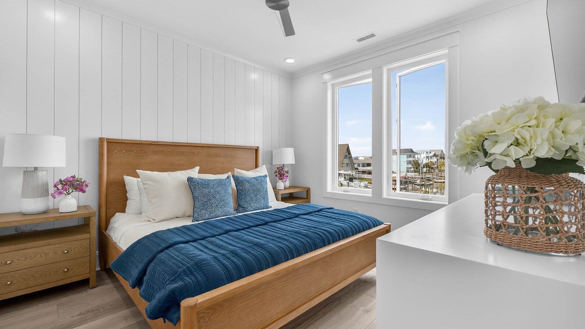 Une chambre à coucher avec un lit en bois, des murs blancs, une table de chevet, une lampe et une vue sur la ville grâce à de grandes fenêtres.