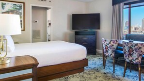 Ein Hotelzimmer mit einem Bett, einem Schreibtisch mit Lampe, einem Fernseher auf einer Kommode und Blick auf die Stadt durch ein Fenster.