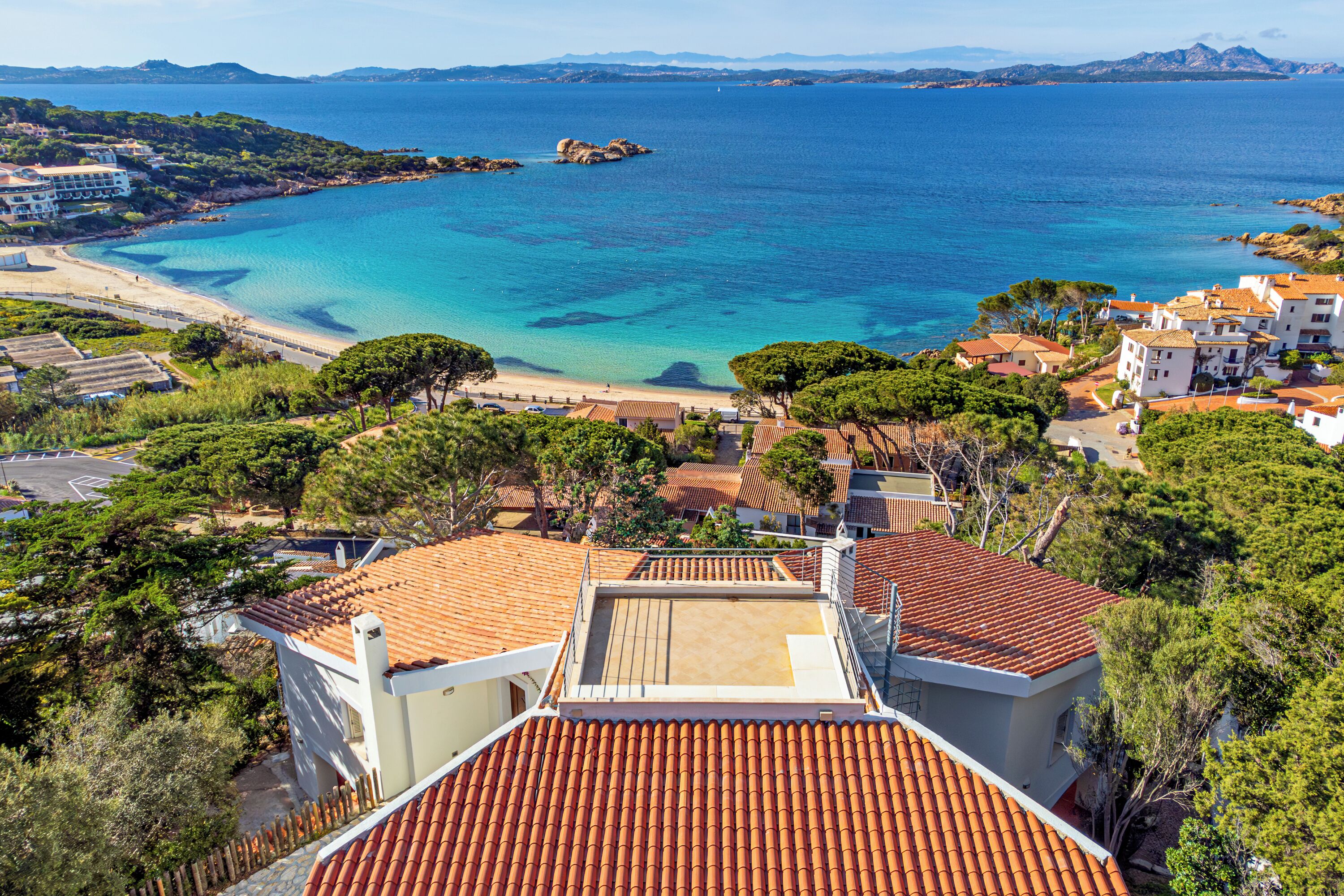 A coastal residential area with terracotta roofs, a beach, and clear blue waters.