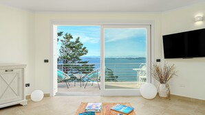 A modern living room with a large sliding glass door leading to a balcony with a view of the sea. There is a wooden coffee table with magazines and a remote control, a white vase with branches, and a flat-screen TV mounted on the wall.