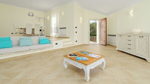 A spacious living room with a wooden coffee table, a sofa with cushions, a dining area with chairs, and a white cabinet.