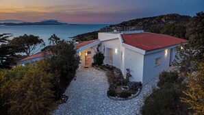 A coastal house with a stone pathway leading to the entrance, surrounded by greenery and overlooking the sea.