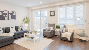 Un salon moderne avec un canapé gris, une table basse blanche et un fauteuil gris. De grandes fenêtres sont équipées de stores, une horloge est accrochée au mur et une plante est posée sur la table basse.