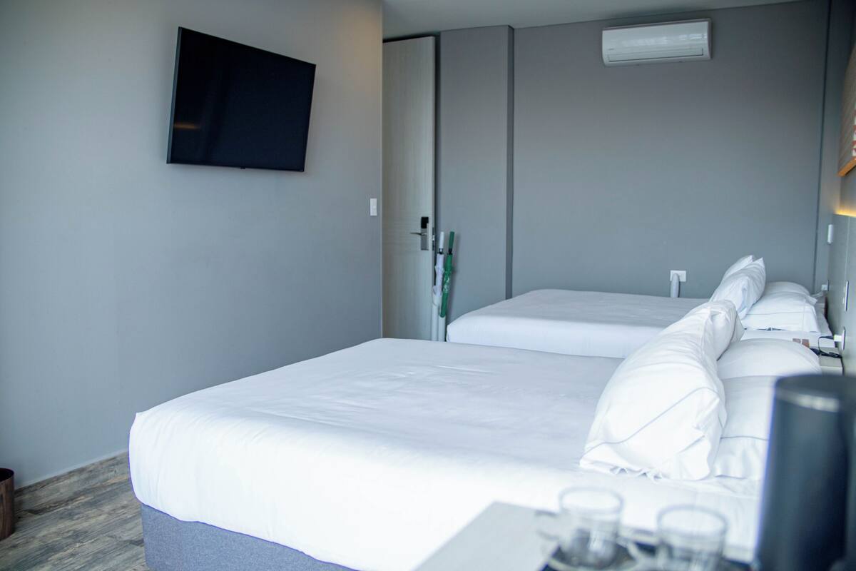 A hotel room with a large bed, a flat-screen TV mounted on the wall, and a bedside table with a lamp and a glass of water.