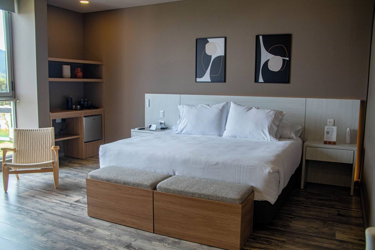 A modern hotel room with a large bed, wooden headboard, and a small seating area. There are two bedside tables with lamps and a shelf with decorative items.