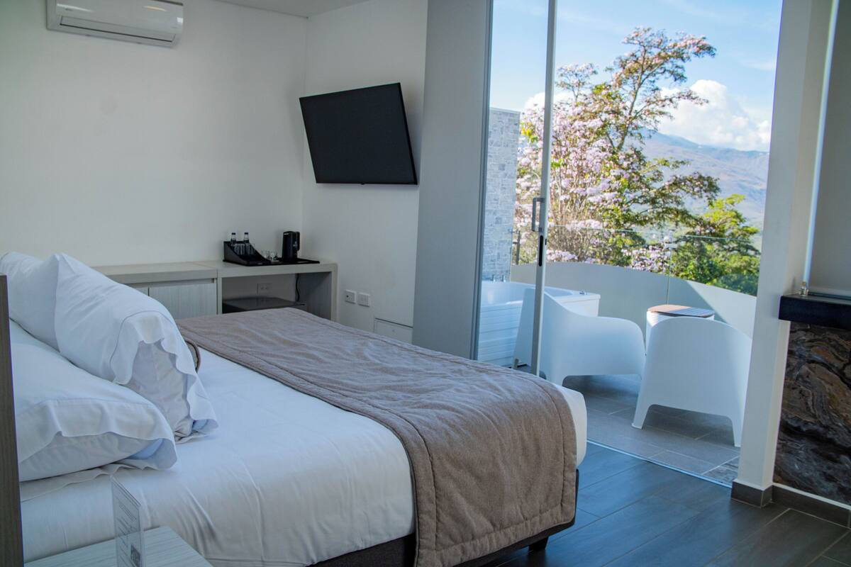 A modern bedroom with a bed, a flat-screen TV, a small desk, and a balcony with a view of trees and mountains.