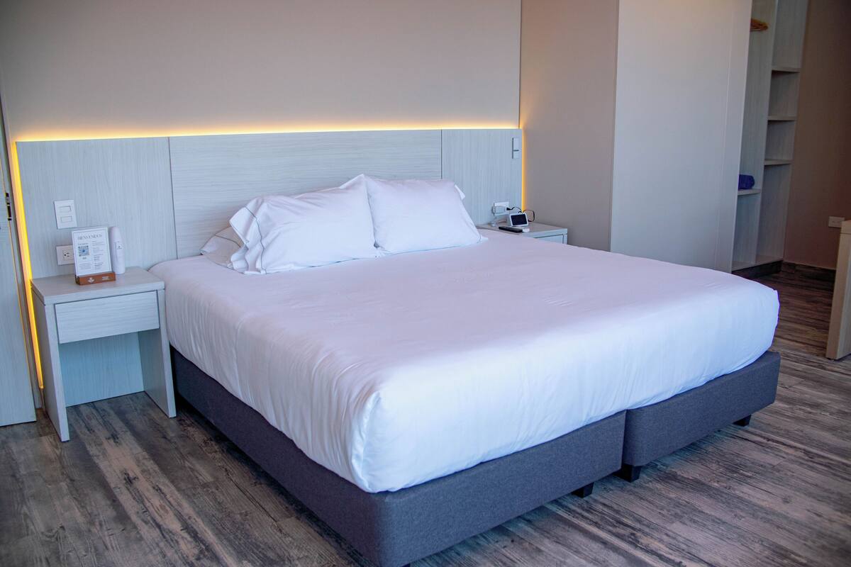A modern hotel room with a large bed, a nightstand with a lamp, and a built-in shelf.