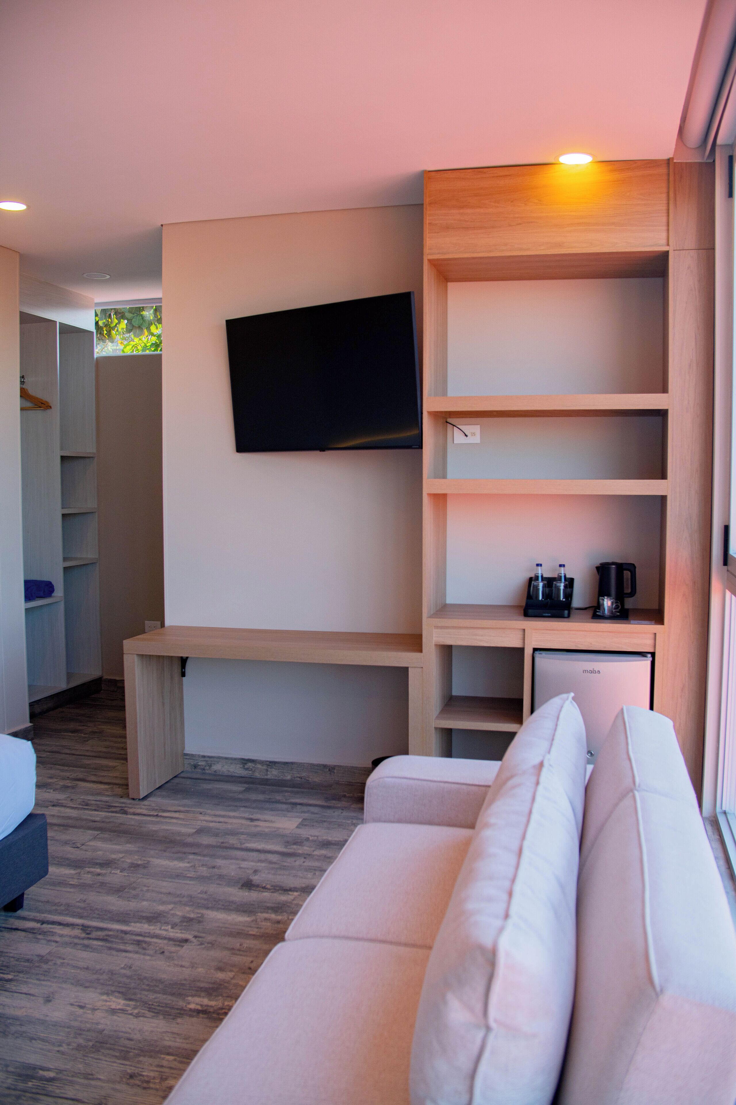 A modern hotel room with a wooden bench, a sofa, a flat-screen TV mounted on the wall, and a minibar.