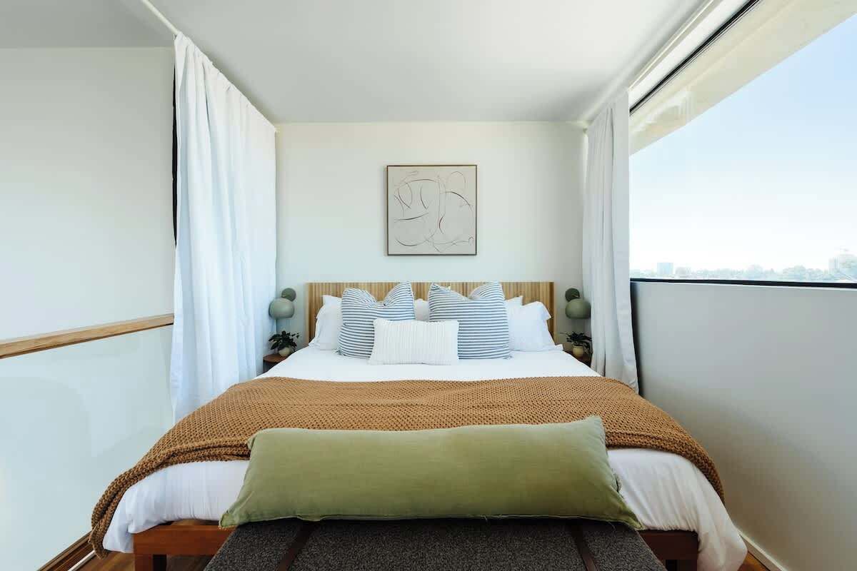 Ein ordentlich bezogenes Bett mit einer braunen Decke, weißen Kissen und einem grünen Kissen. Darüber an der Wand hängt ein gerahmtes abstraktes Kunstwerk.