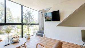 A modern living room with a large window, a flat-screen TV mounted on the wall, a round wooden table, and a sofa. - Hillcrest Hideaway – Stylish Loft in the Heart of SD (San Diego)