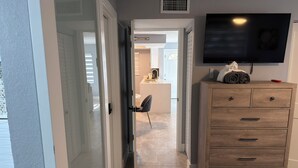 Corredor que dá para uma cozinha com área de refeições, uma televisão montada na parede e um aparador de madeira com gavetas.