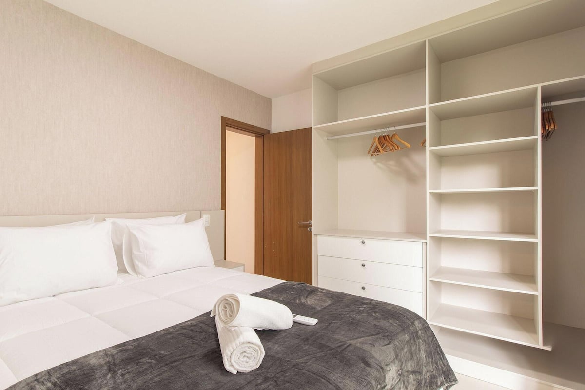 A hotel room with a bed, a grey blanket, a rolled white towel, and a white wardrobe with hanging space.