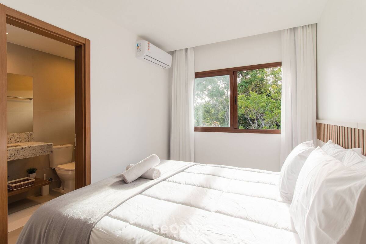 A bedroom with a bed, a window with curtains, a bathroom with a toilet, and an air conditioning unit.
