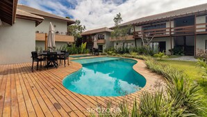 A swimming pool with a wooden deck, surrounded by greenery and a two-story building with balconies. - Houses in Gated Community With Pool and BBQ Area - PCR (Porto Seguro)