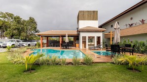 A pool area with a covered seating area, lounge chairs, and a barbecue grill. - Houses in Gated Community With Pool and BBQ Area - PCR (Porto Seguro)