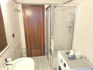 A bathroom with a wooden door, a shower with a glass door, a sink, and a small shelf with toiletries. - Città di Brescia adorable 4 person apartment with AC in Brescia Hospital Area (Brescia)
