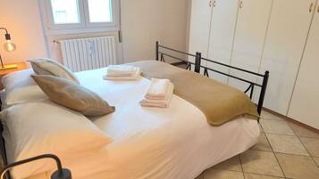 Een opgemaakt bed met kussens en gevouwen handdoeken, een bedlamp, een radiator, en een inloopkast met schuifdeuren.