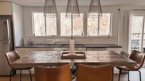 Une cuisine moderne avec une grande table en bois, quatre chaises marron et des luminaires suspendus.