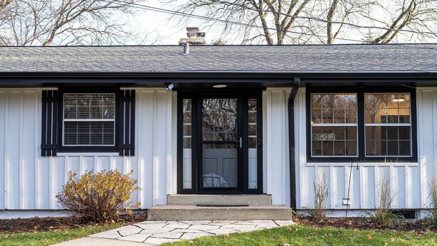 Ett envåningshus med en veranda på framsidan, en svart dörr och en liten trädgård framför huset, med en gångstig som leder till entrén.