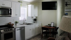 Eine kompakte Küche mit weißen Schränken, einer Mikrowelle, einem Backofen und einem Geschirrspüler. Es gibt einen kleinen Essbereich mit Tisch und Stühlen sowie einen an der Wand befestigten Fernseher.