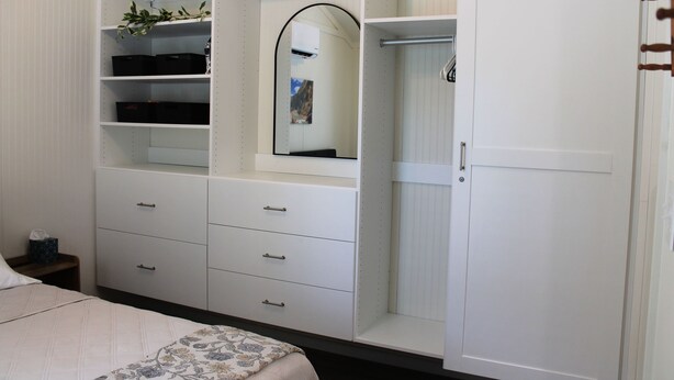Ein Schlafzimmer mit einem Bett, einem Schrank mit zahlreichen Fächern, einem Spiegel und einem kleinen Nachttisch.