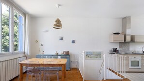Une cuisine moderne avec une table et des chaises en bois, un luminaire suspendu et une vue sur l’extérieur grâce à de grandes fenêtres.